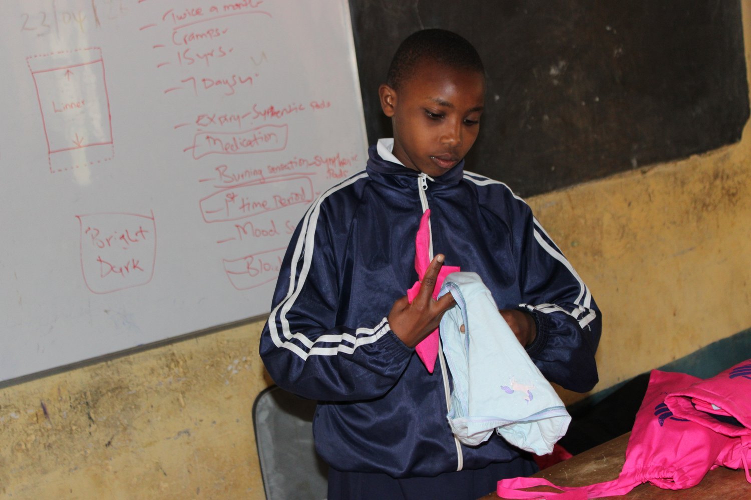A pupil in a navy tracksuit demonstrates how to use a reusable menstrual kit in front of a whiteboard of health notes.