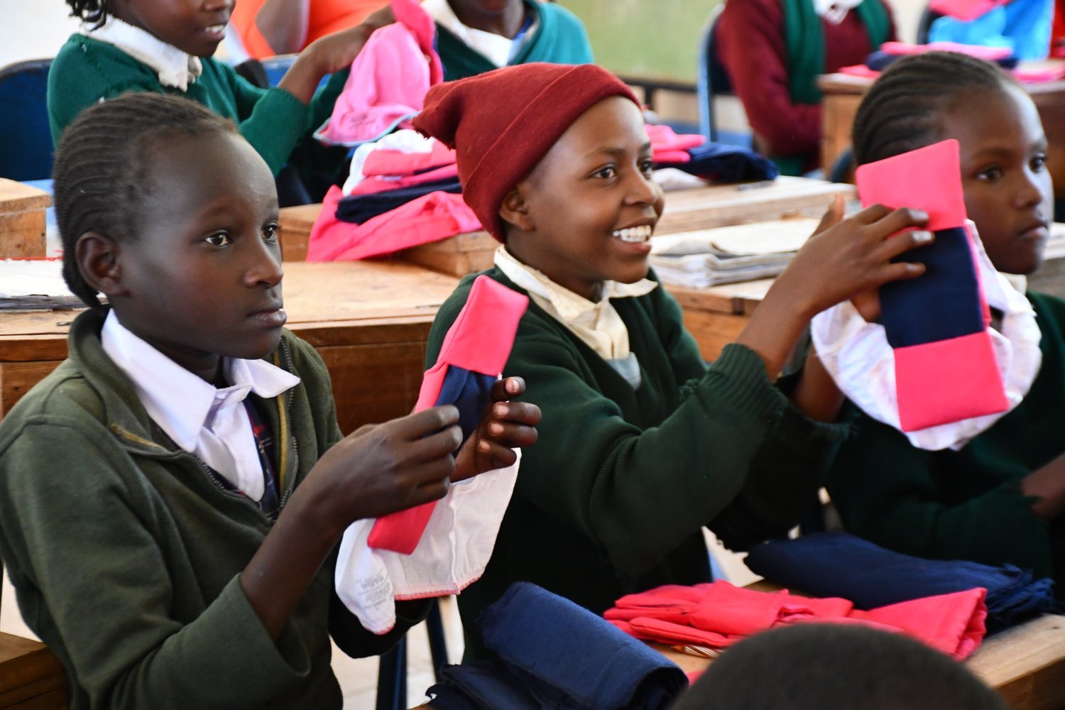 Girls in green school uniforms holding pink and navy fabric items at wooden desks in a classroom.