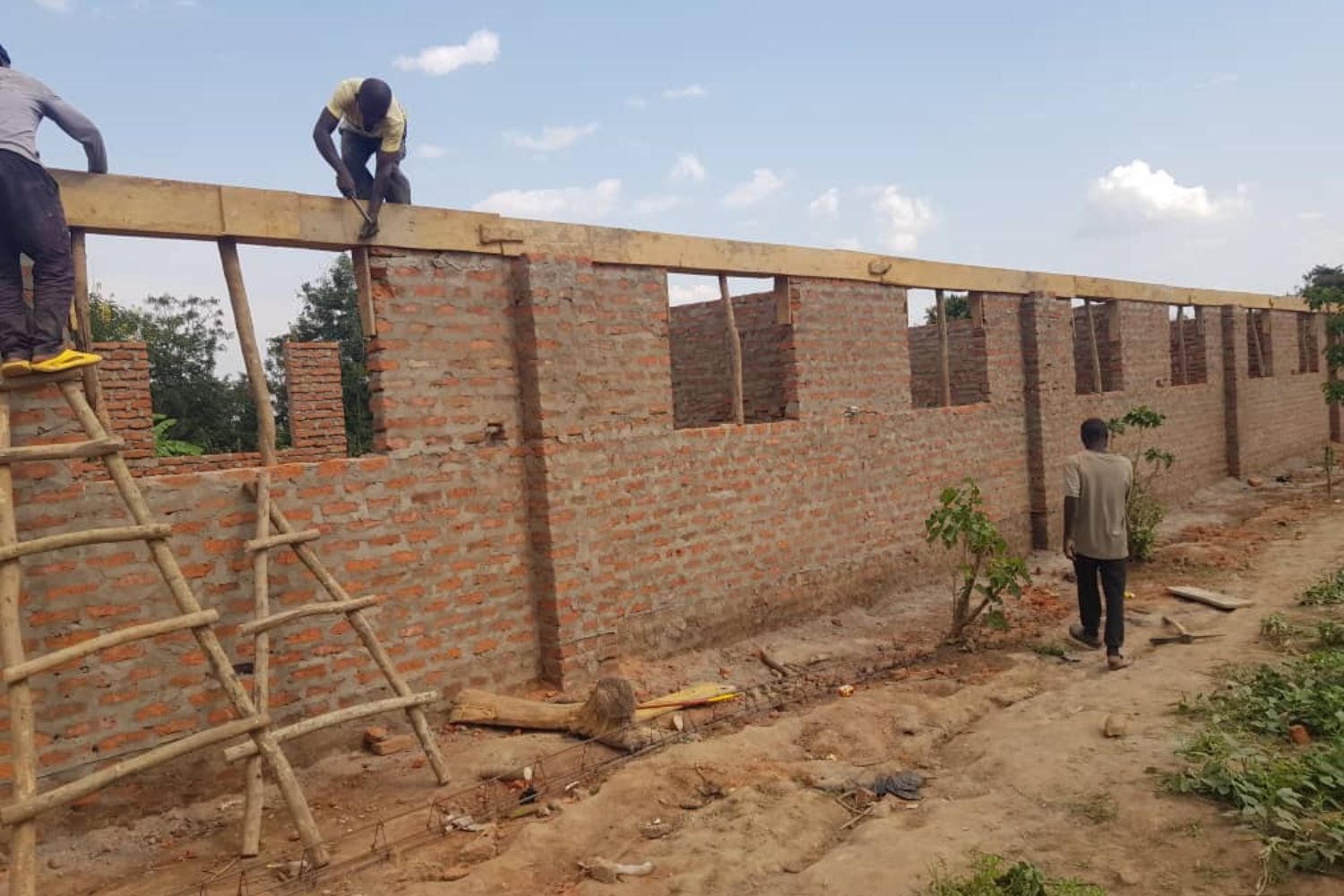 Workers constructing a brick building with wooden scaffolding under a clear sky. One man is on the scaffolding, another on the ground.