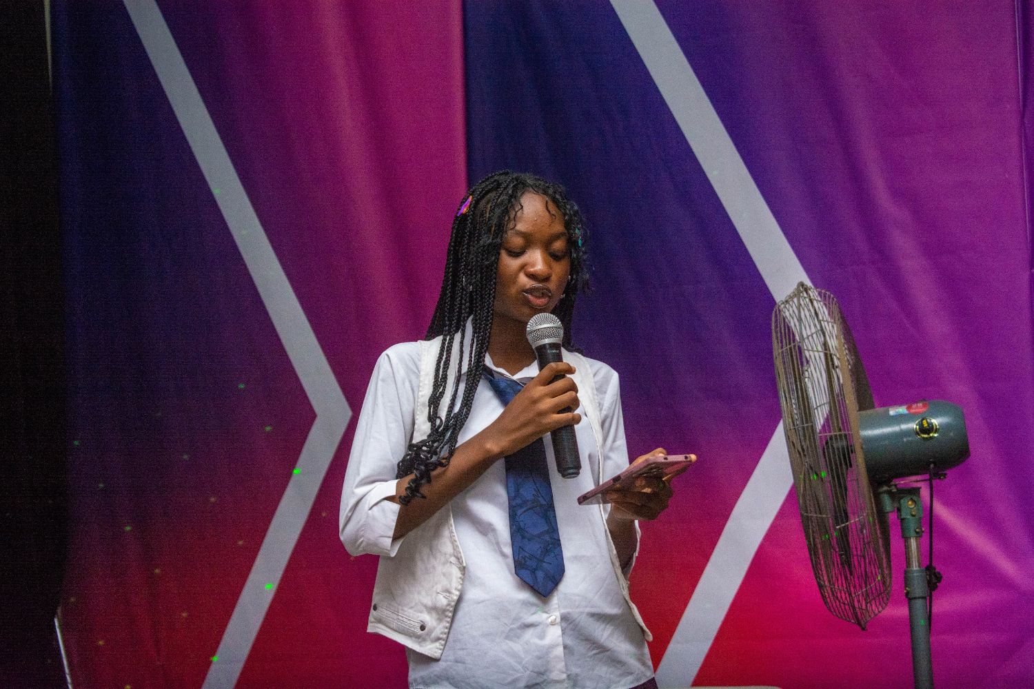 Young woman speaks into a microphone, holding a phone, on a stage with a vibrant purple and pink backdrop, and a nearby standing fan.