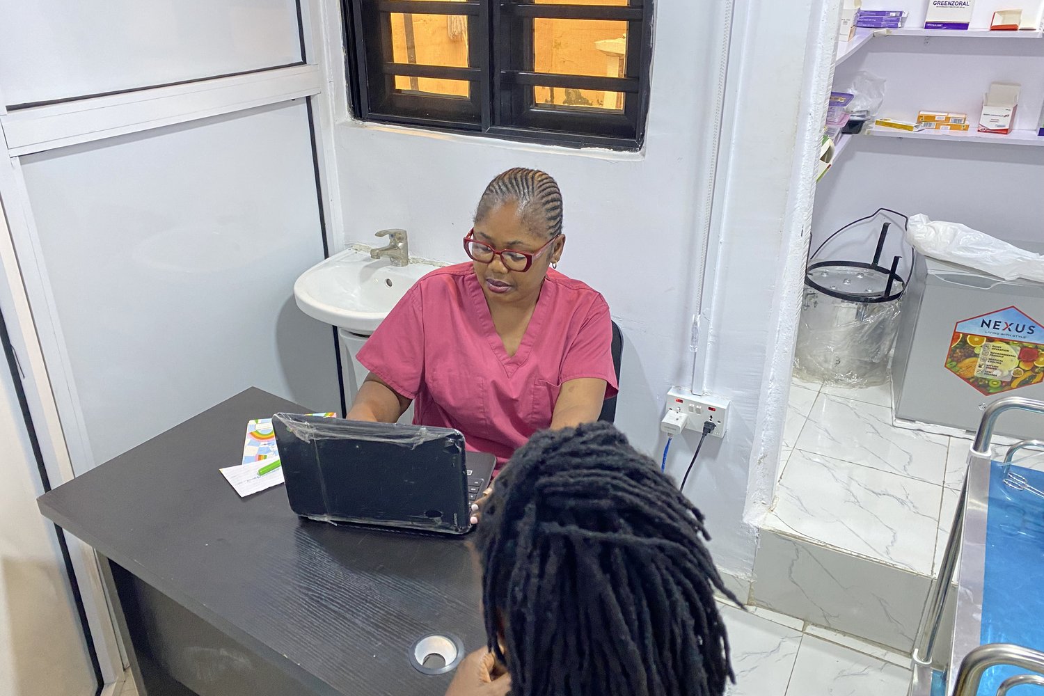 A healthcare worker in pink scrubs consulting with a patient at a desk with a laptop in a medical clinic.