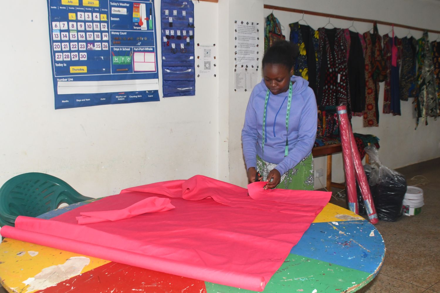 A person in a purple hoodie cuts bright pink fabric on a colorful table in a room with patterned clothing and a schedule on the wall.