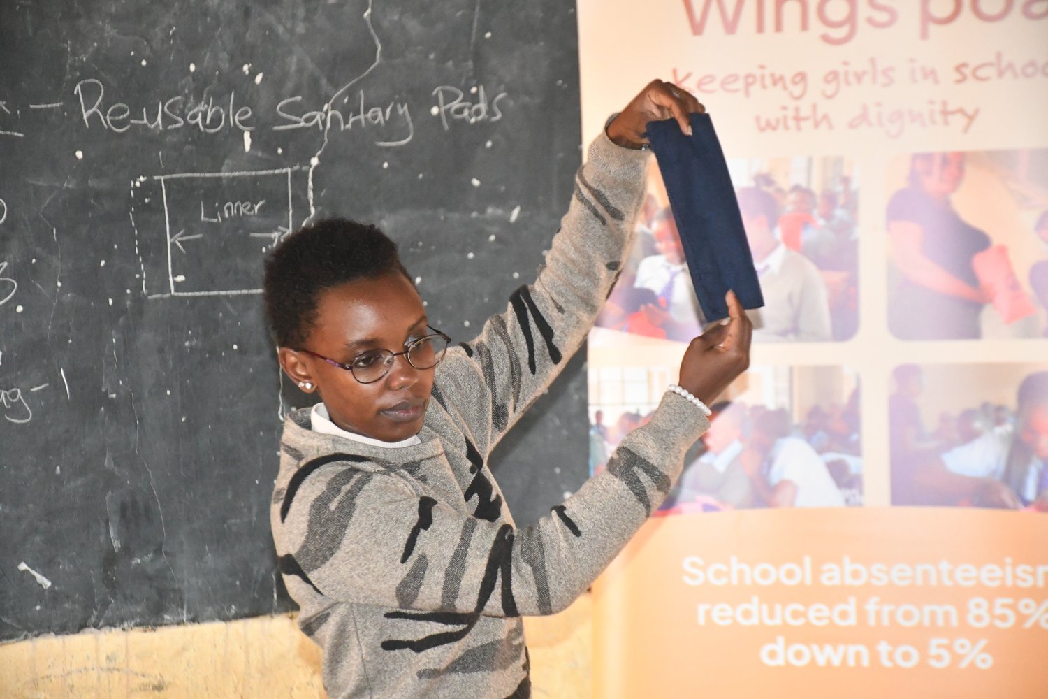 A woman in a grey patterned sweatshirt holds up a dark blue reusable sanitary pad beside a chalkboard in a classroom.