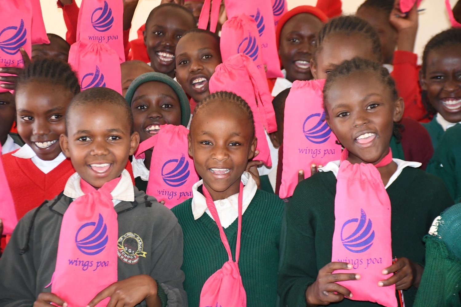 Children in green and red uniforms holding pink "Wings Poa" branded drawstring bags in a crowded indoor setting.