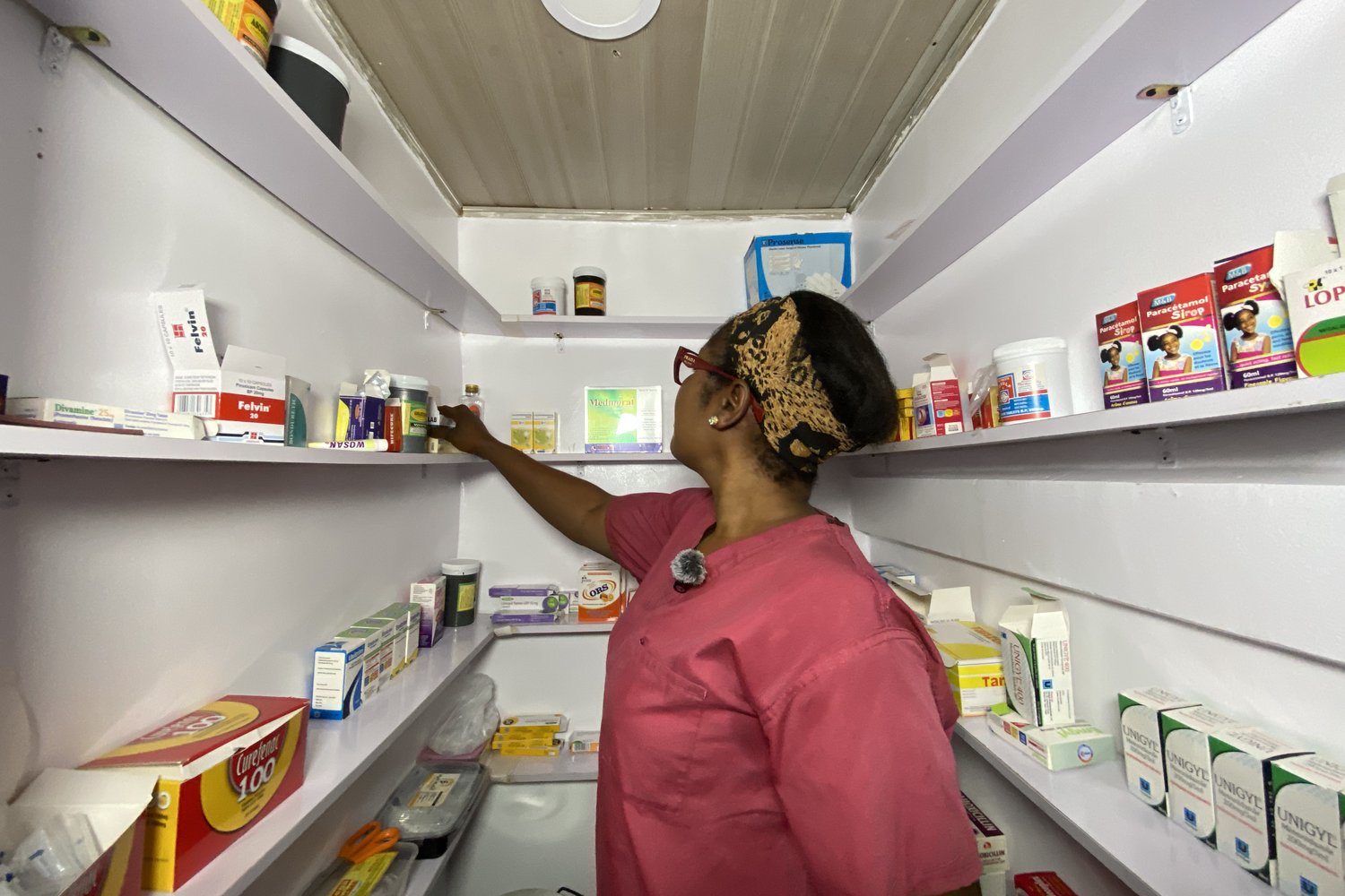 A woman in a pink uniform reaching for medicine on shelves stocked with pharmaceutical products in a small pharmacy.