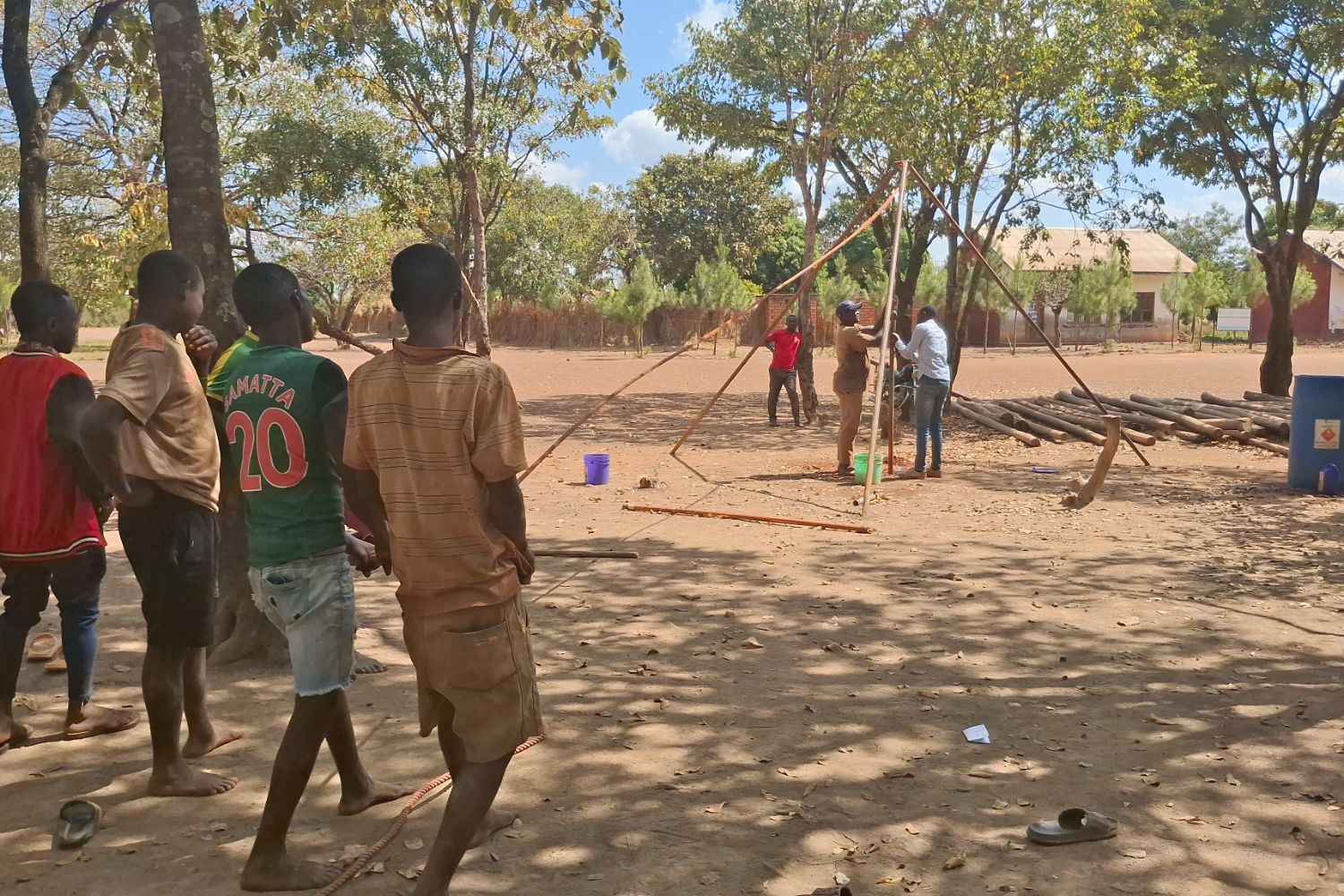 People gather outdoors, watching a group setting up a wooden structure. Trees and buildings are in the background under a clear sky.