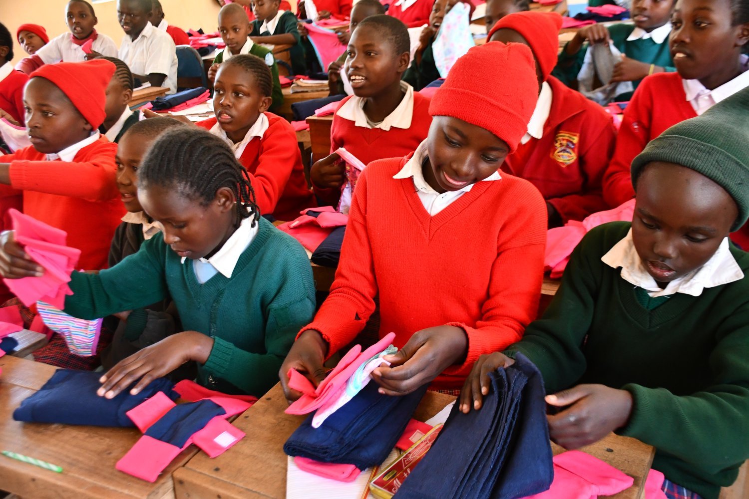 Children in red and green school uniforms examining pink and navy fabric items at wooden desks in a classroom.