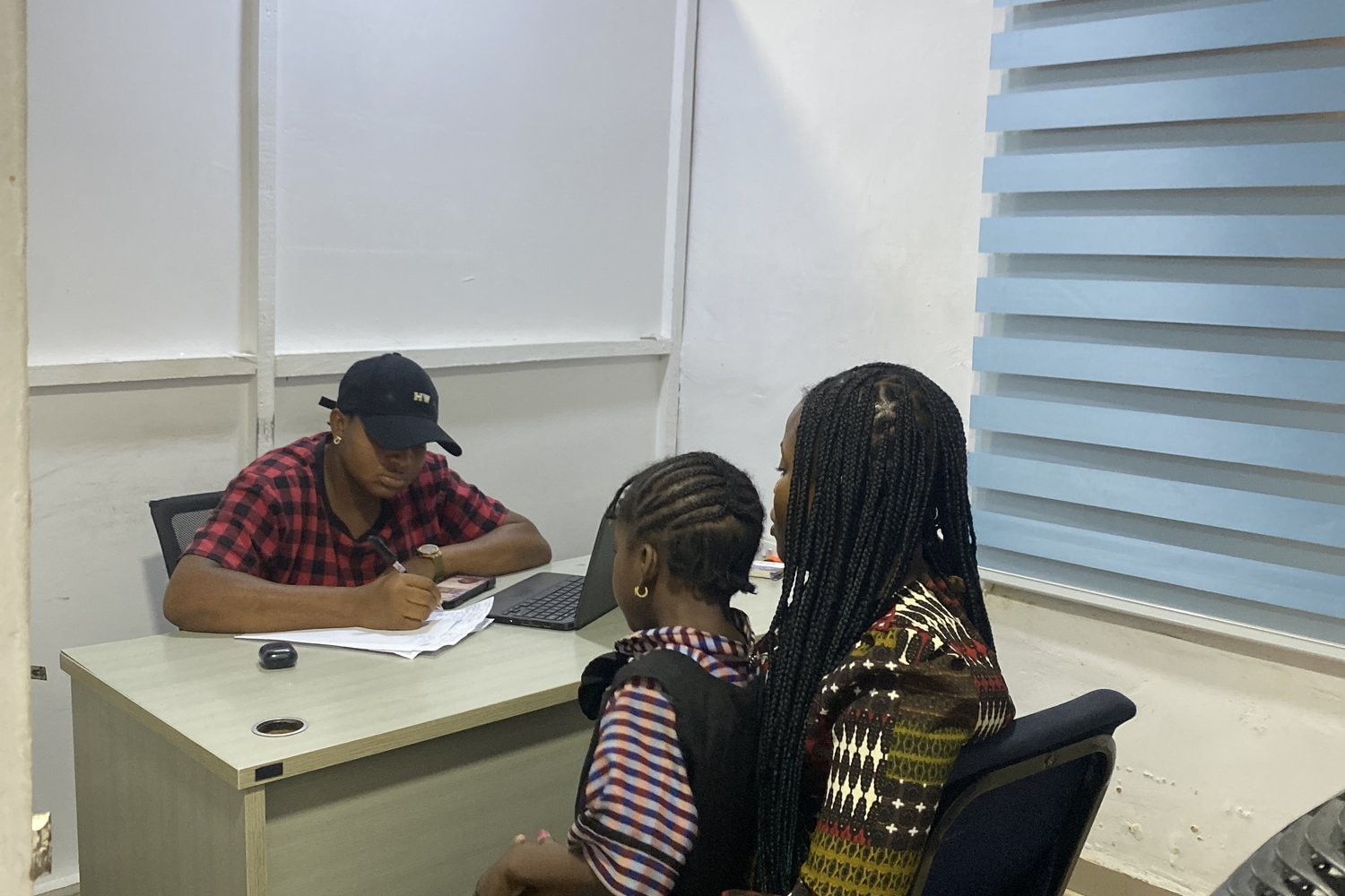 Three people gathered around a desk with a laptop in an indoor office, one seated wearing a red checked shirt and cap.