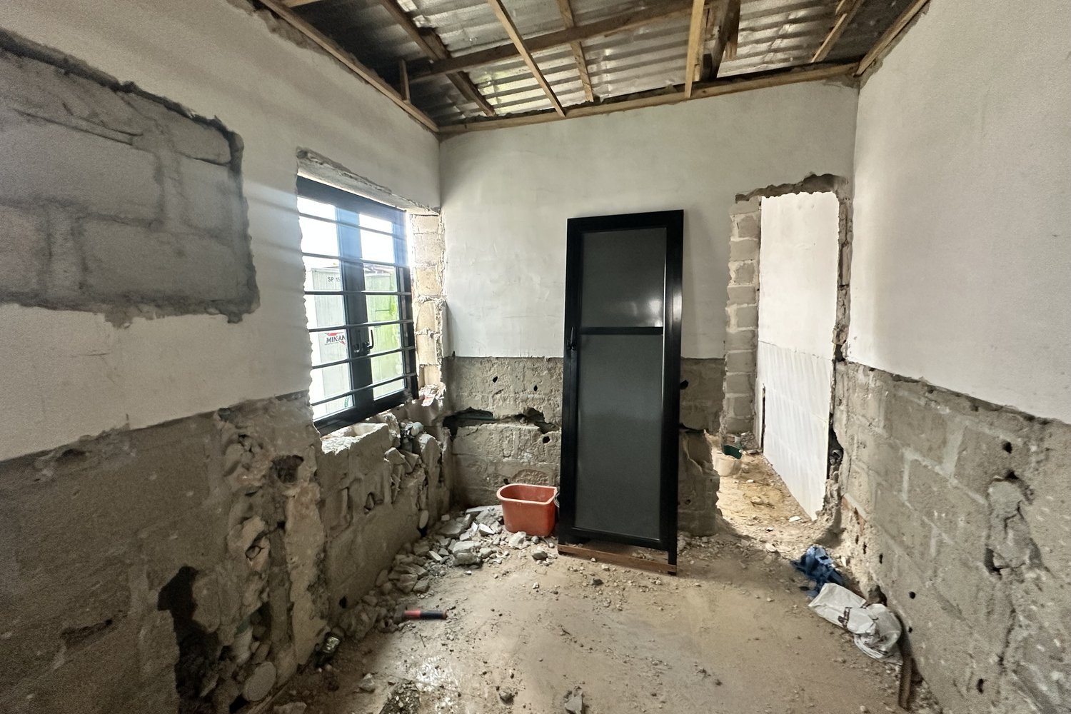 A black door leaning against a partially plastered wall in a room undergoing renovation with exposed brickwork and debris on the floor.