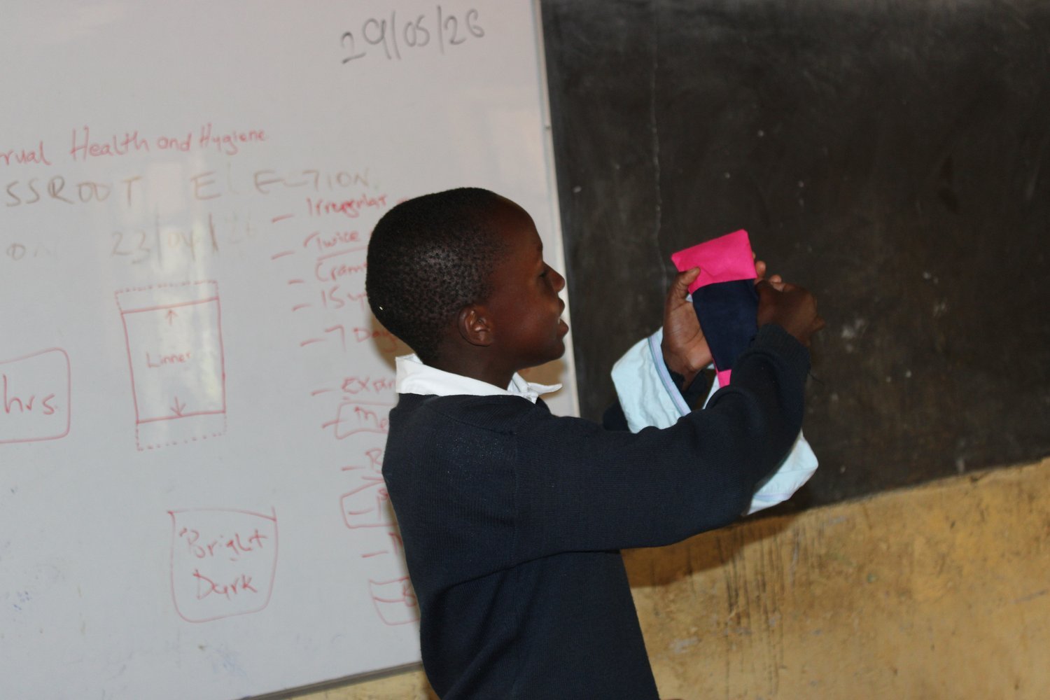 A child in a dark navy school uniform holds a pink menstrual cup in front of a whiteboard displaying health and hygiene notes.