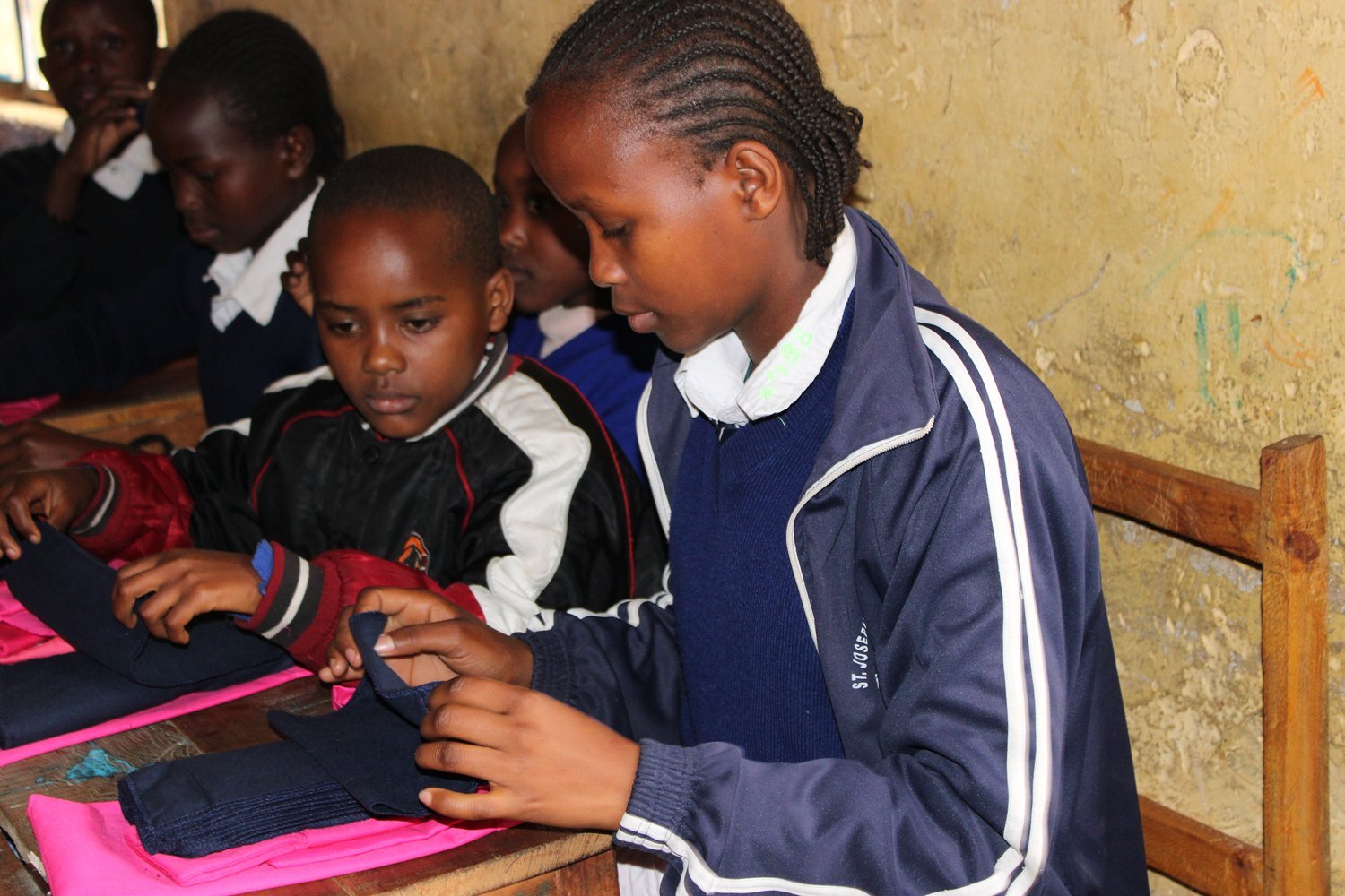 Children in blue school uniforms examining a piece of dark blue fabric at a wooden desk in a classroom.