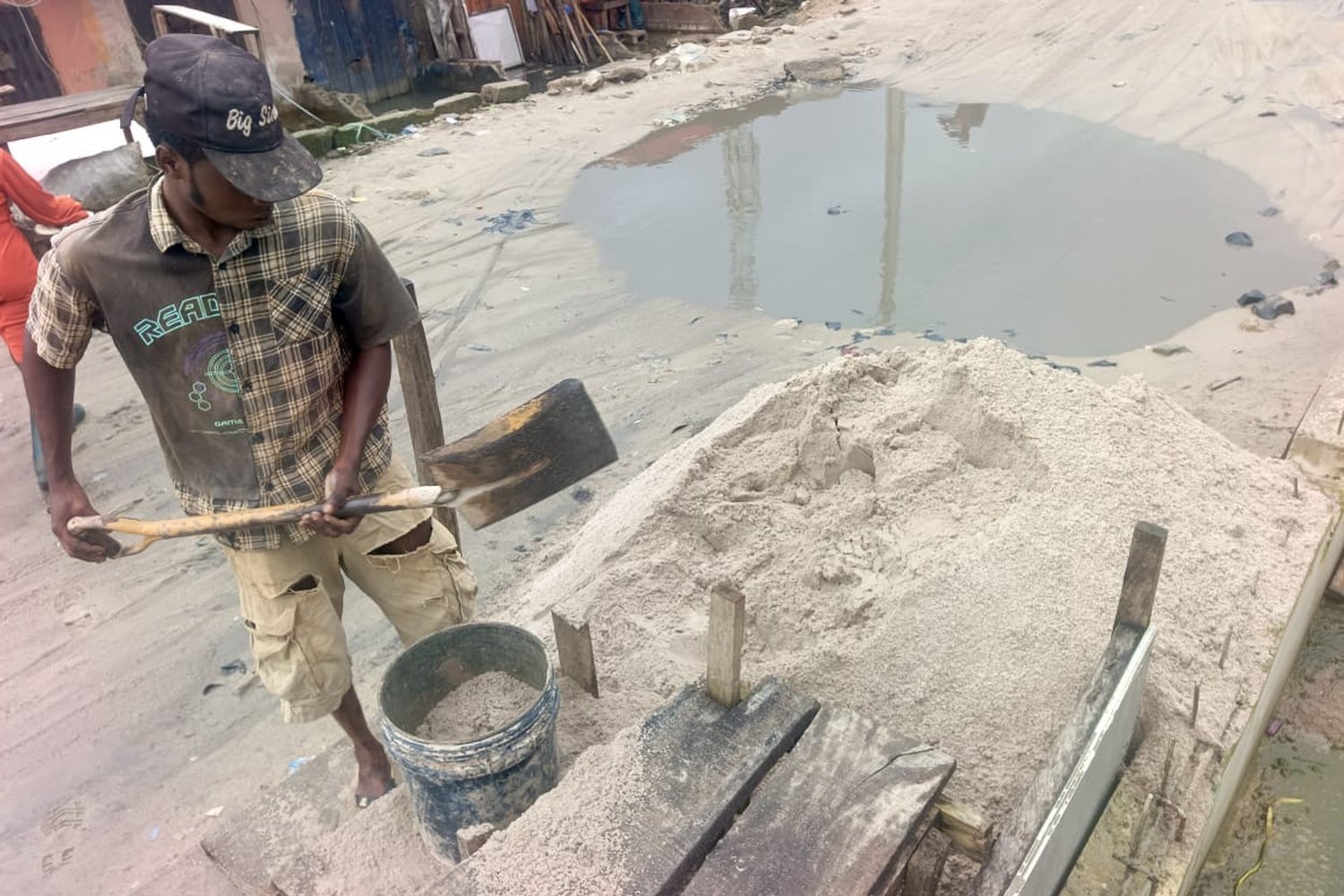 A man in a plaid shirt and cap shovelling sand into a bucket at an outdoor construction site near a puddle.