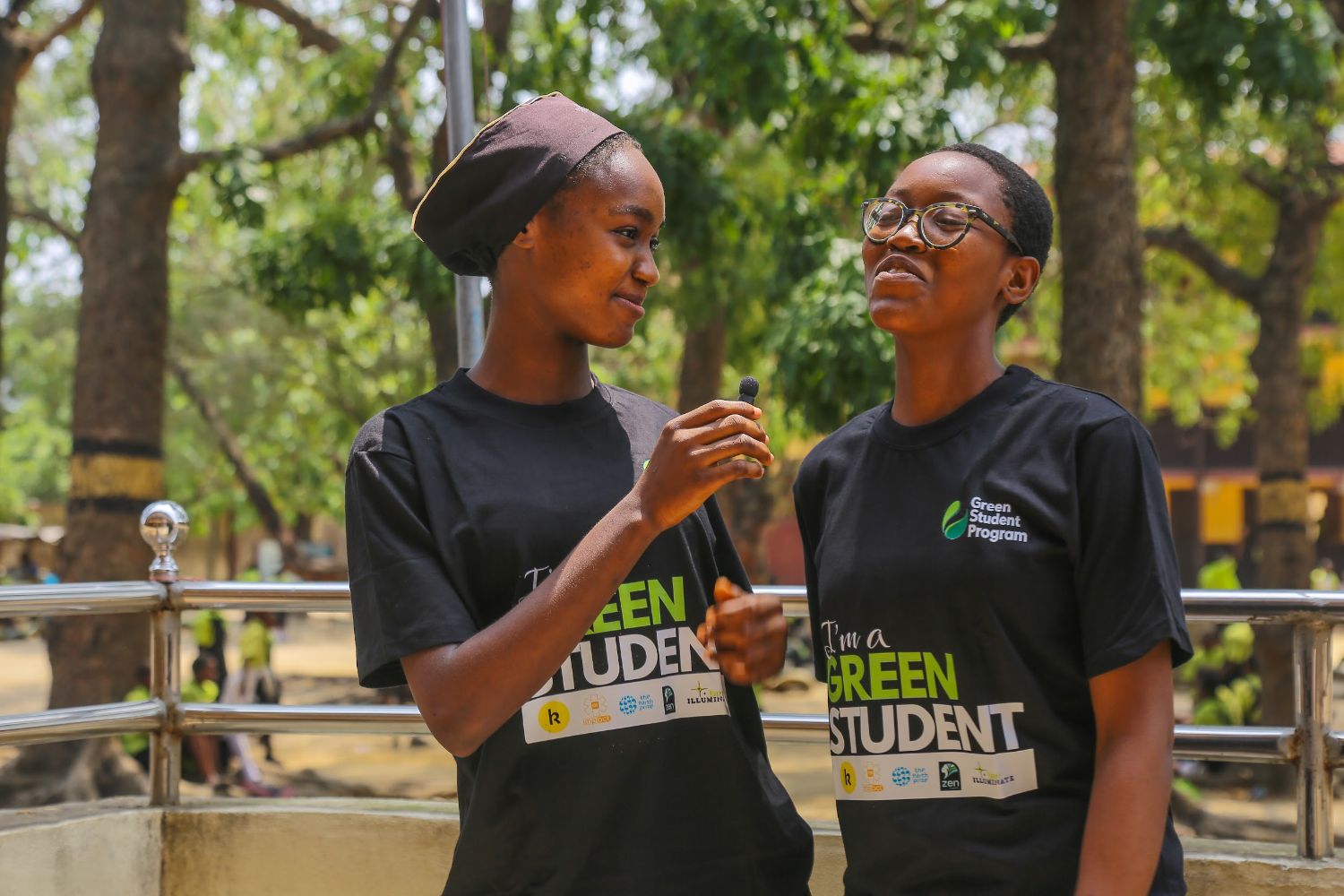 Two people wearing "Green Student Program" shirts stand outdoors, smiling. One holds an ice cream cone. Trees and a railing are in the background.