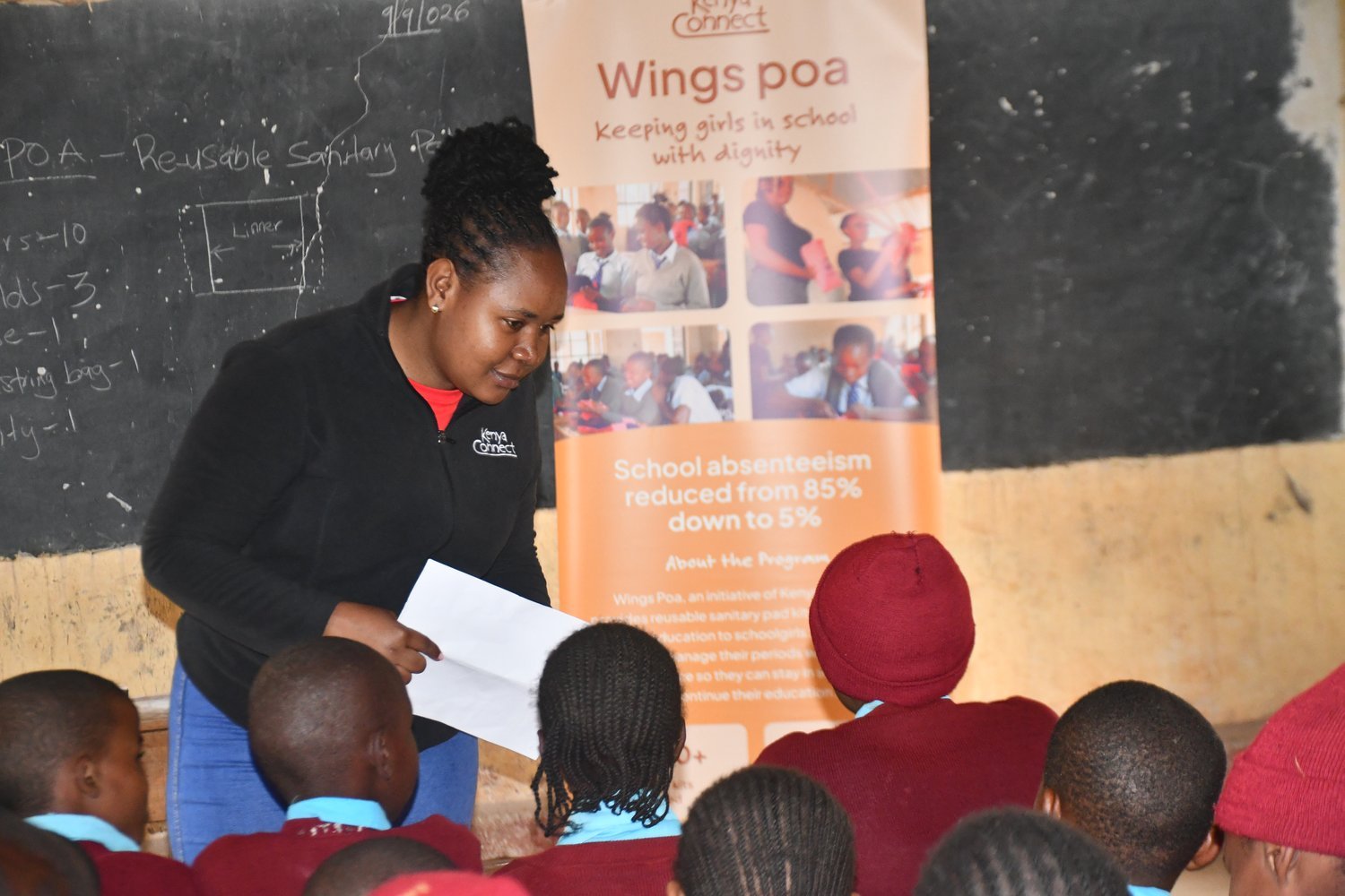 A Kenya Connect presenter holding papers addresses schoolchildren in a classroom beside a "Wings Poa" reusable sanitary pad programme banner.