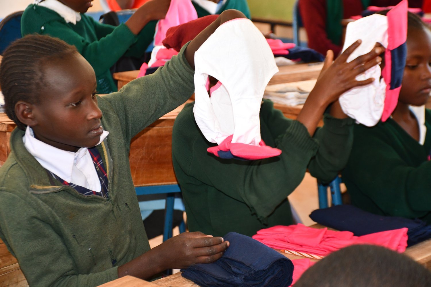 Children in green school uniforms examining clothing items, including underwear, at wooden desks in a classroom.