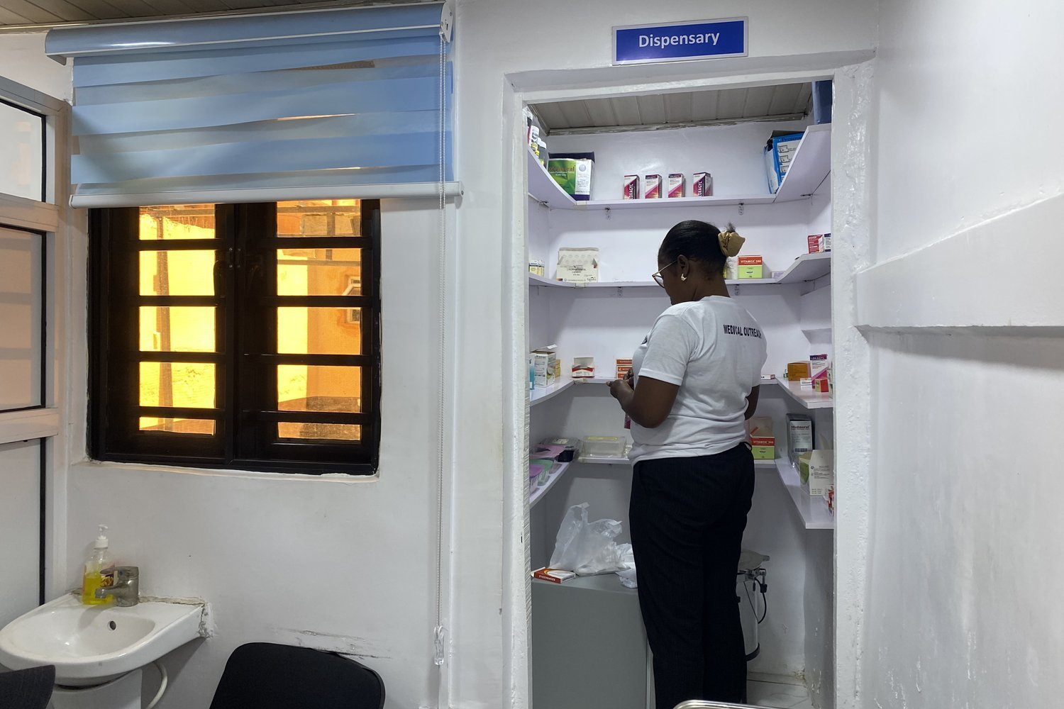 A healthcare worker in a white uniform retrieving medicines from shelves inside a small dispensary room.