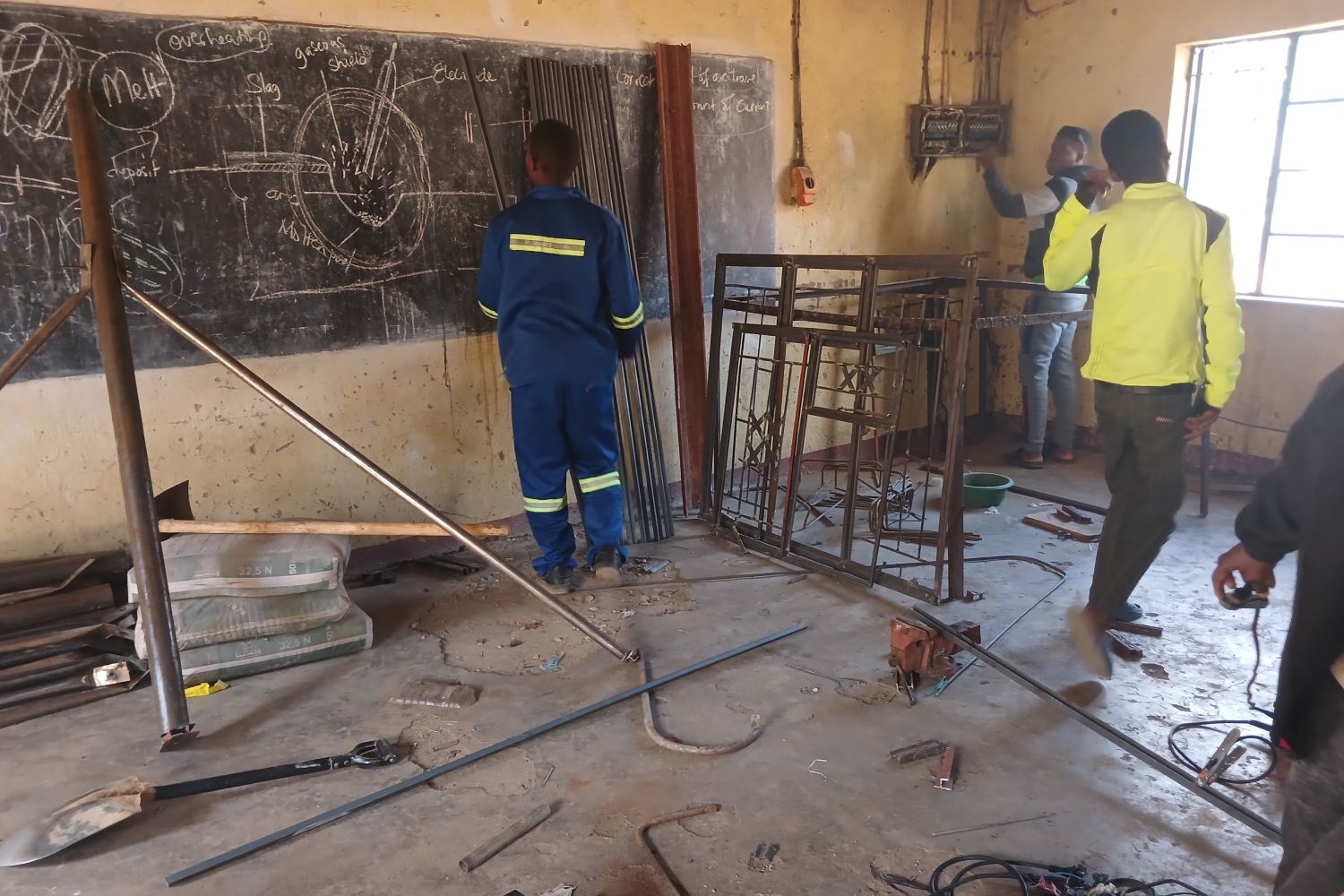 Workers in a workshop with metal bars, tools, and chalkboard diagrams. One man wears a blue uniform, another a bright yellow jacket.