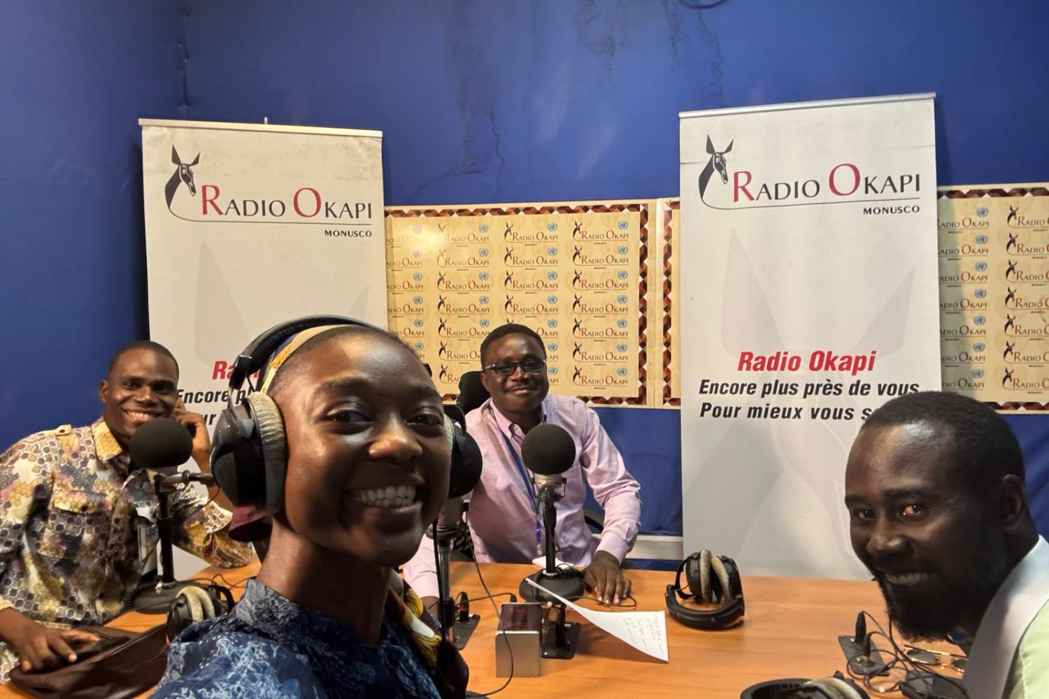 Four people in a radio studio with microphones and Radio Okapi banners, smiling at the camera.