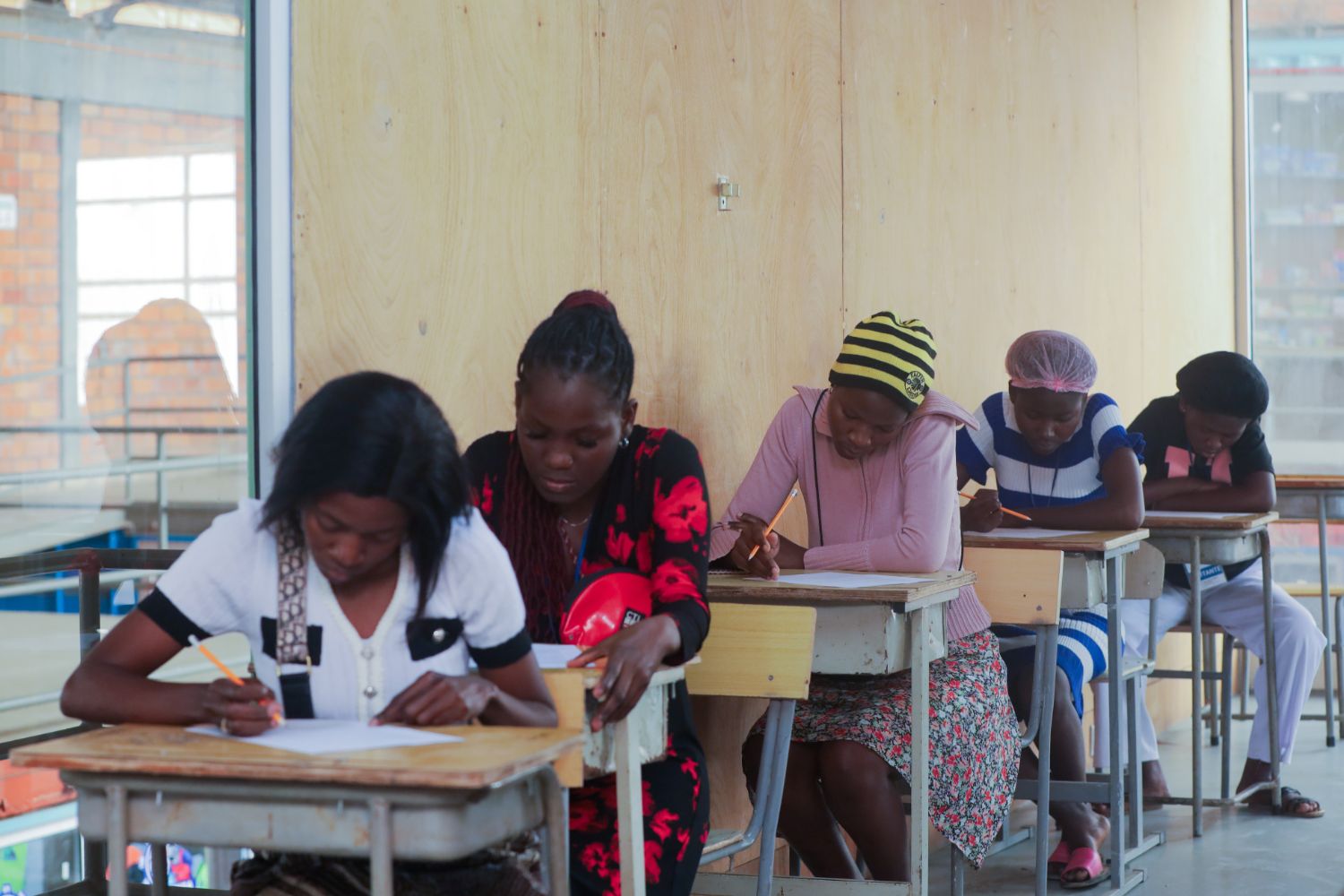 Students sitting at desks in a classroom, focused on writing during an exam. The room has wooden walls and large windows.