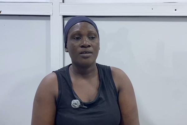 Woman wearing a dark grey headband and black sleeveless top with a lapel microphone, seated in front of a white wall.