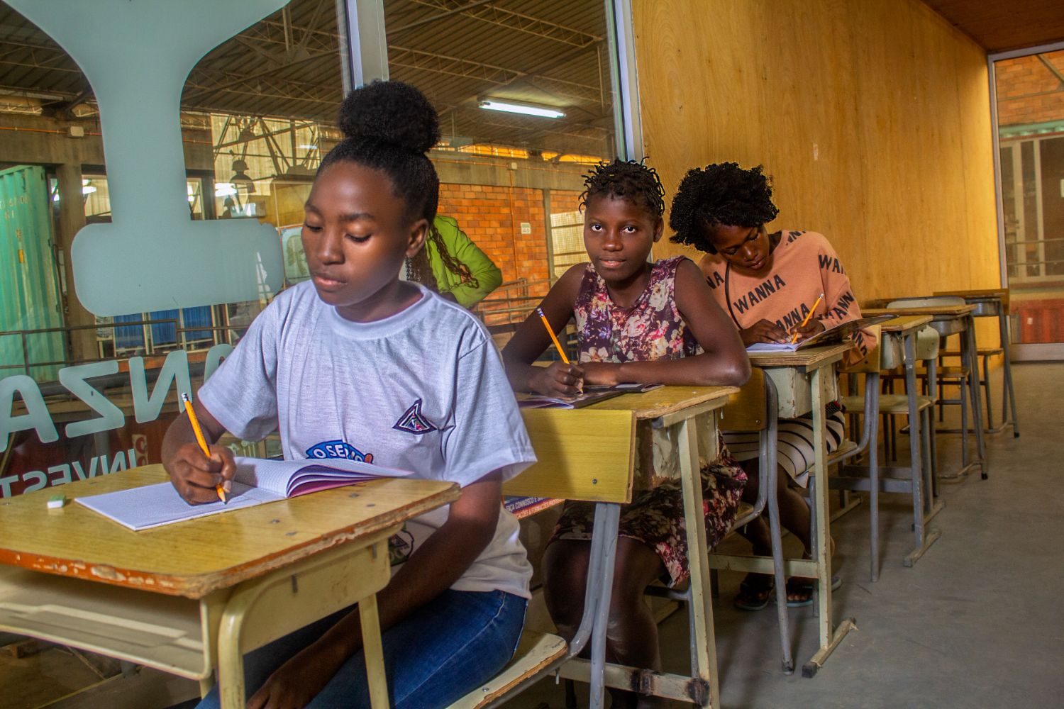 Four students sit in a classroom, writing in notebooks at wooden desks. The room has a mix of brick and glass walls.
