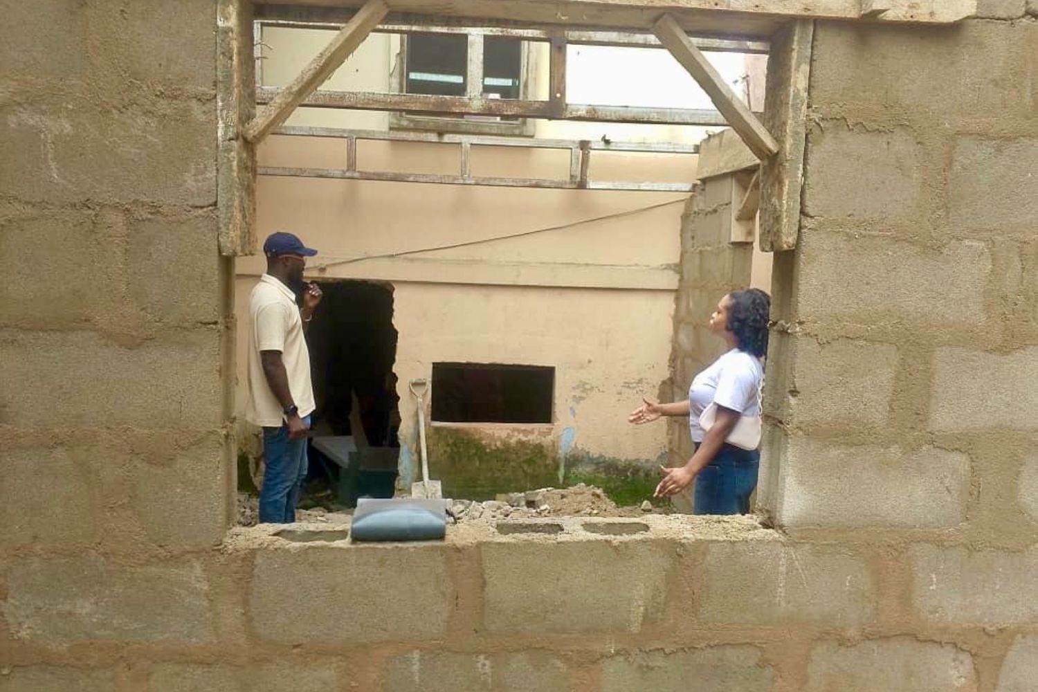 Two people stand in a partially constructed building, conversing through a large window opening in a brick wall.