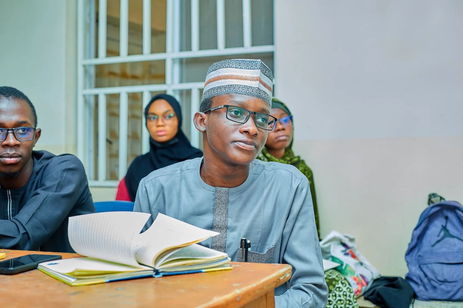 A man in traditional attire sits attentively in a classroom, with an open notebook in front of him. Others are seated behind him.