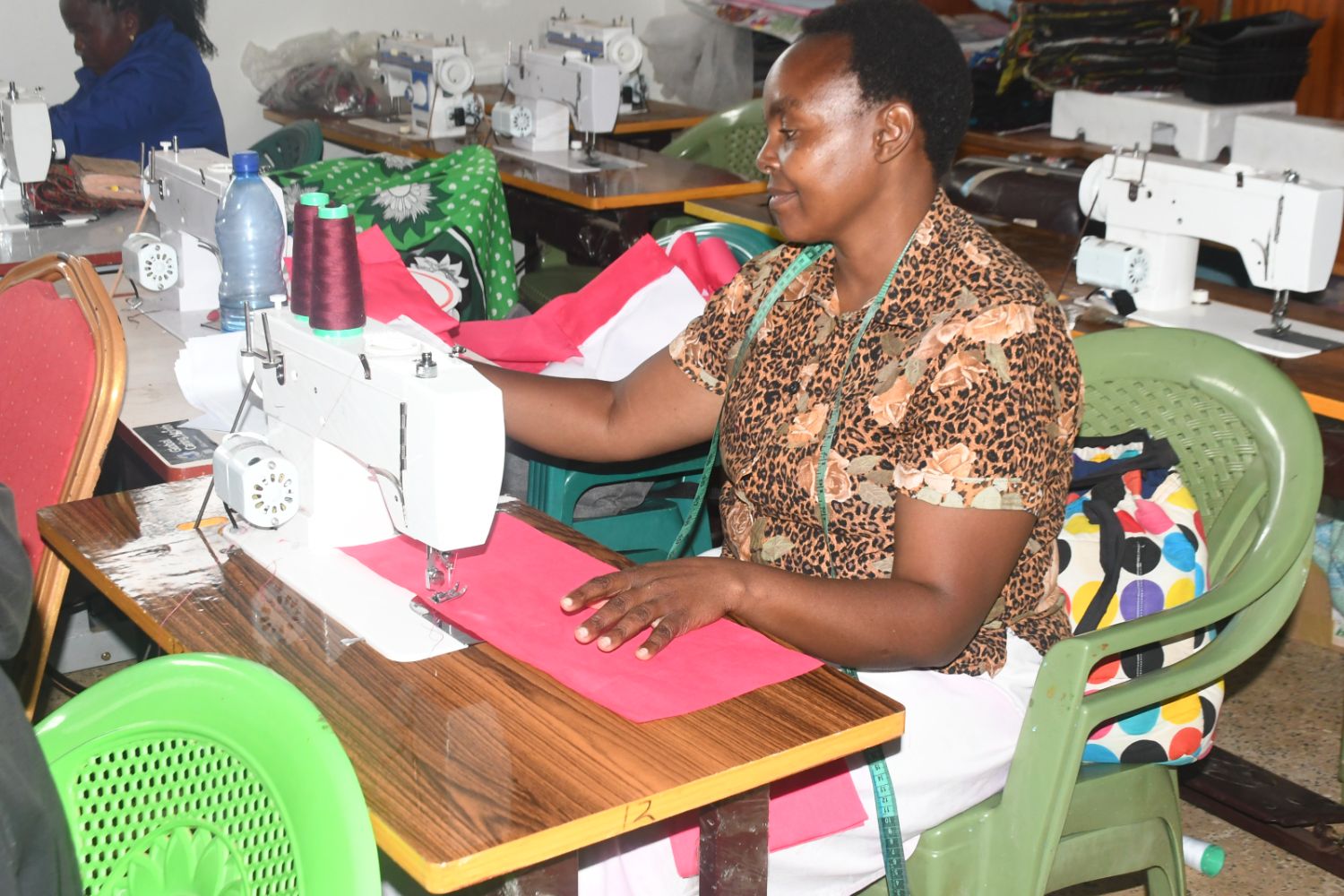 A woman in a patterned shirt sews red fabric on a sewing machine in a room with multiple machines and colorful materials.
