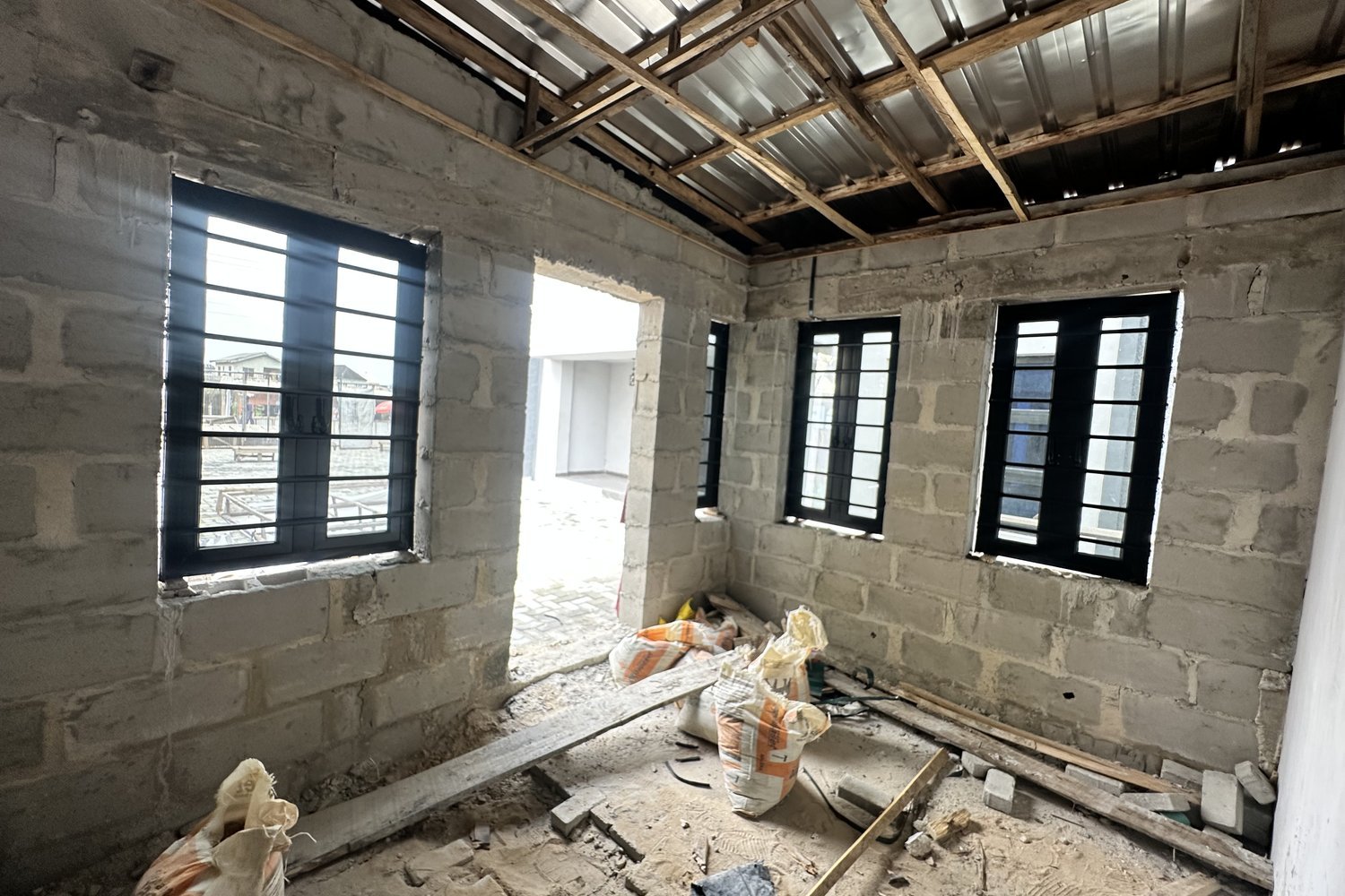 Interior of an unfinished concrete block building under construction, with black-framed windows, exposed timber roof structure, and building materials scattered on the floor.