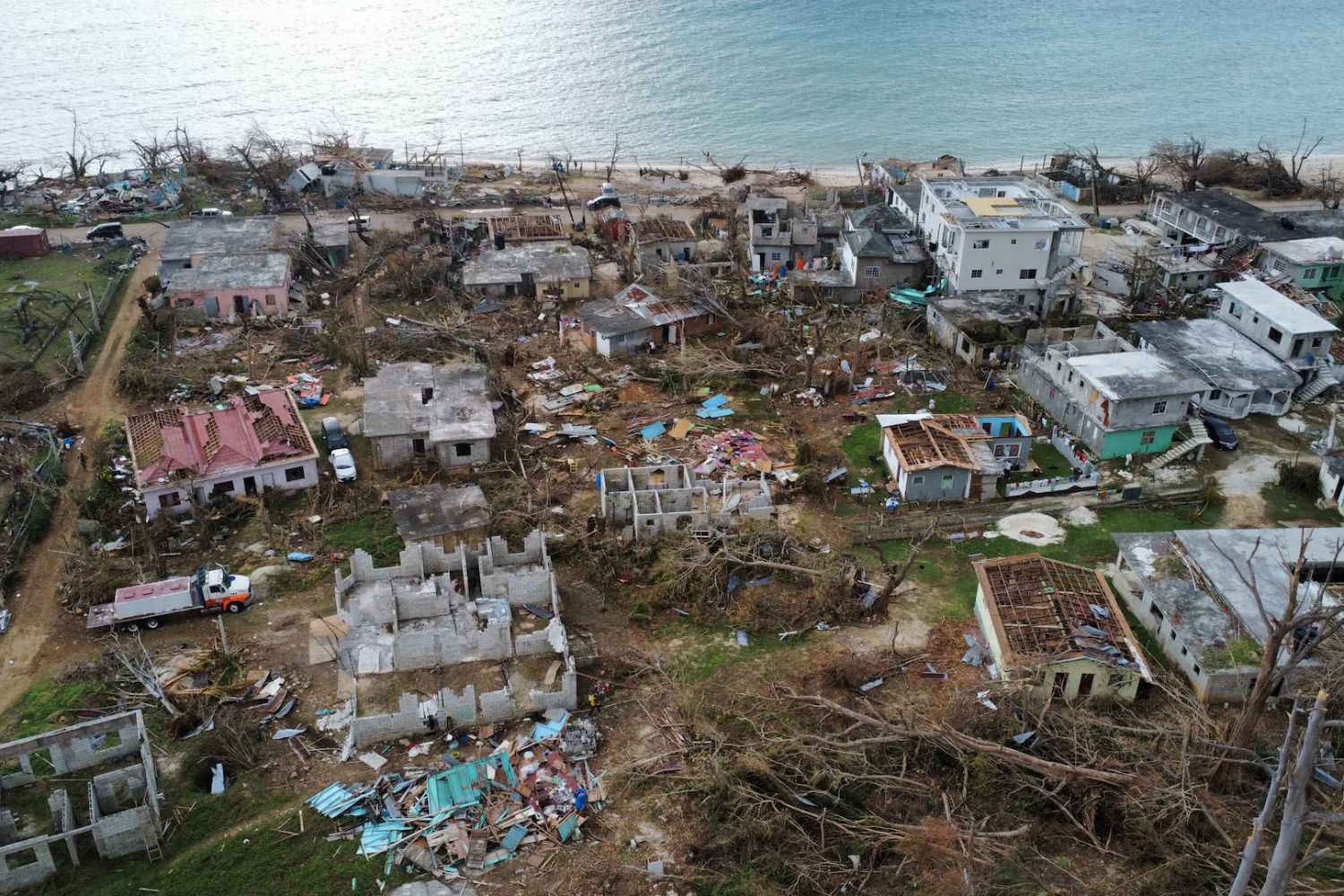 Aerial view of a coastal town devastated by a storm, with damaged buildings, debris, and uprooted trees near the shoreline.