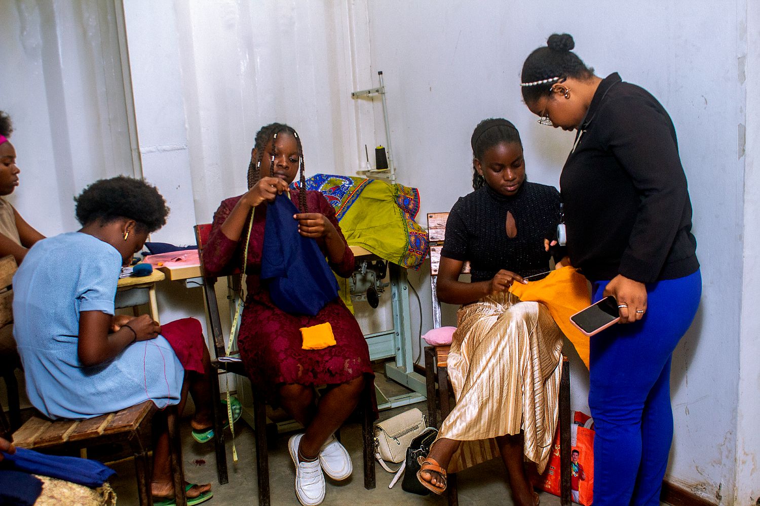 A group of women engaged in sewing, with one woman instructing others. They are seated and focused on their tasks in a small room.