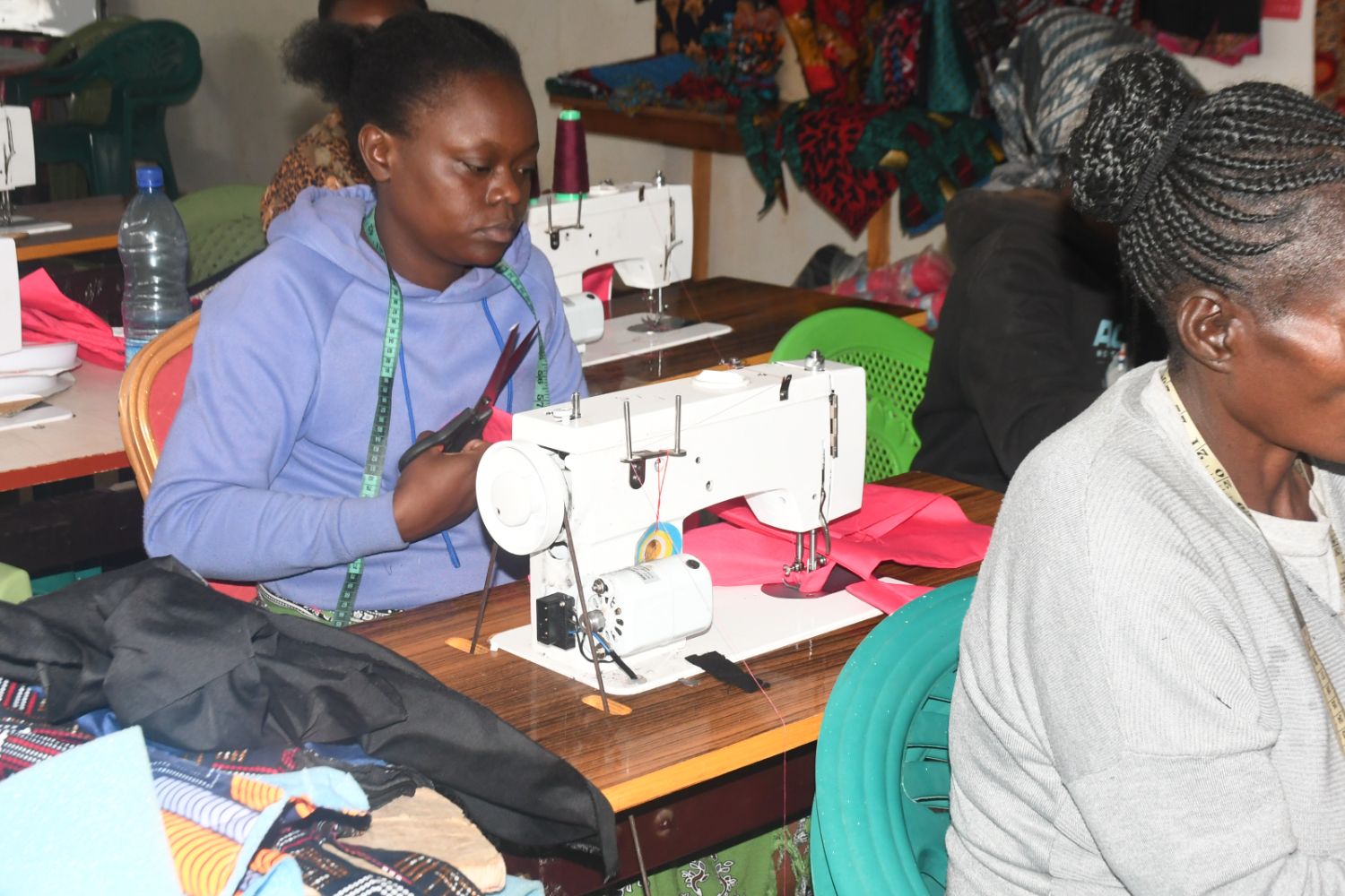 Women sitting at sewing machines, working with fabric in a workshop setting. One woman is holding scissors.