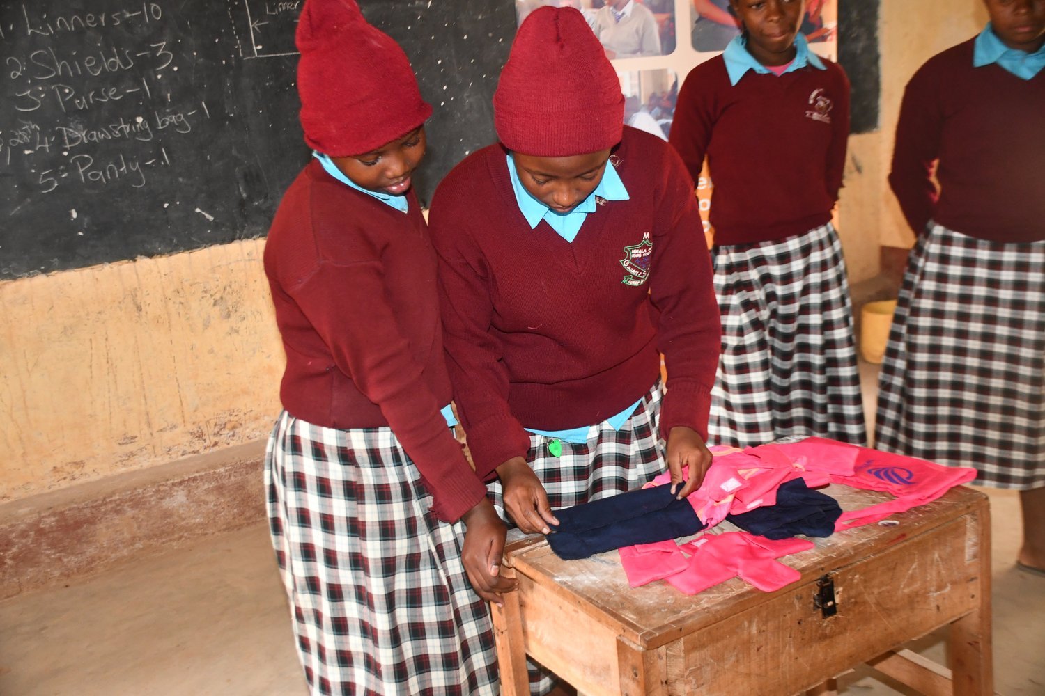 Girls in maroon school uniforms examining clothing items on a wooden desk in a classroom with a chalkboard.