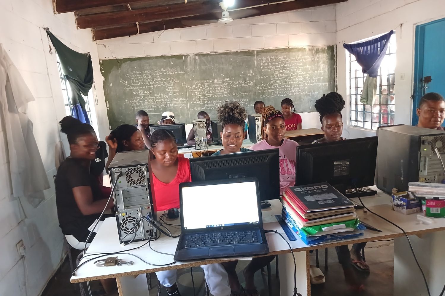 A group of people in a classroom working on desktop computers, with a chalkboard and various equipment visible in the background.