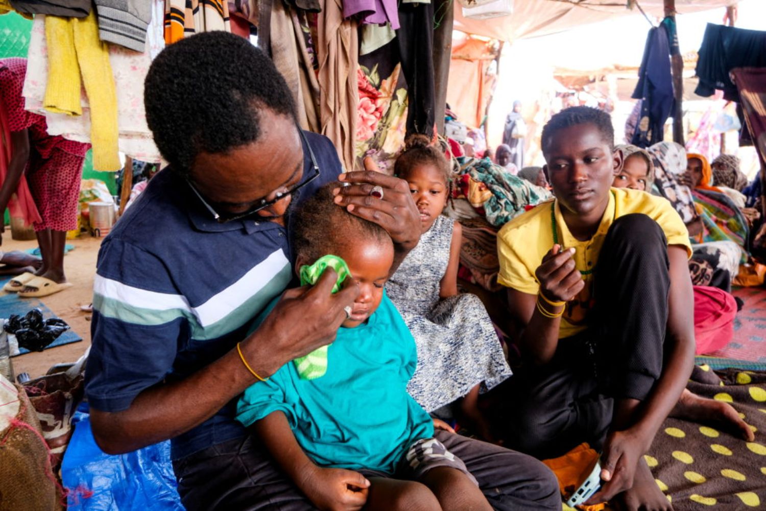 A man gently cleans a young child's face with a cloth in a crowded room, surrounded by several people, including a seated woman and other children.