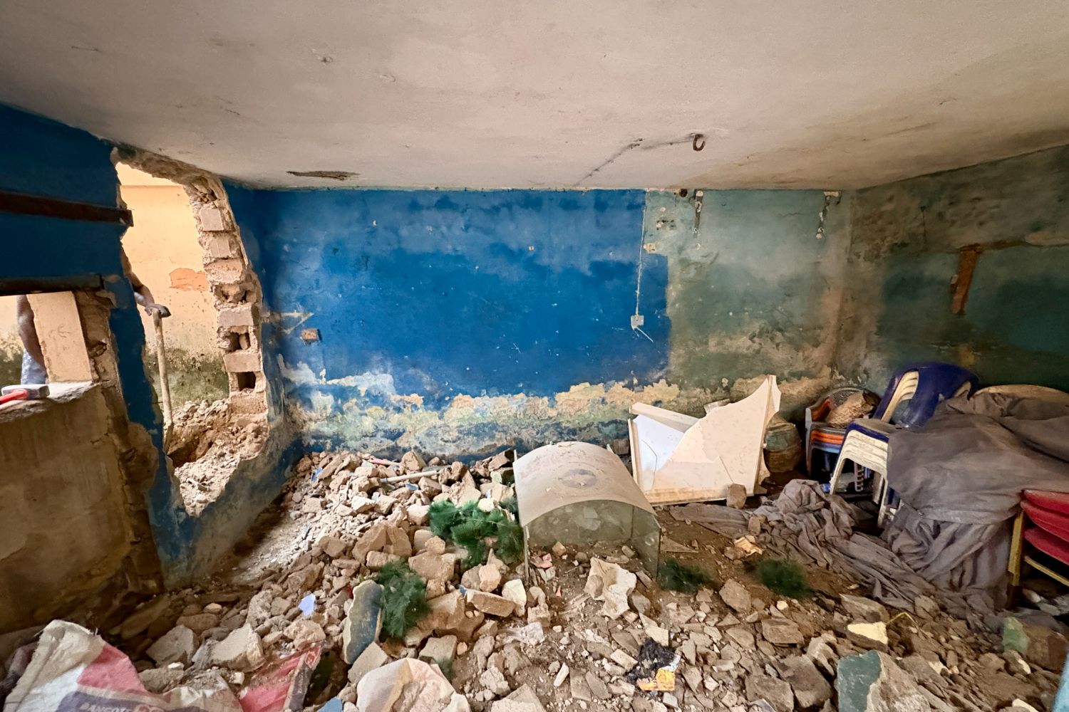 Room with blue walls, debris, and a large hole in the wall. Broken furniture and scattered items are visible on the floor.