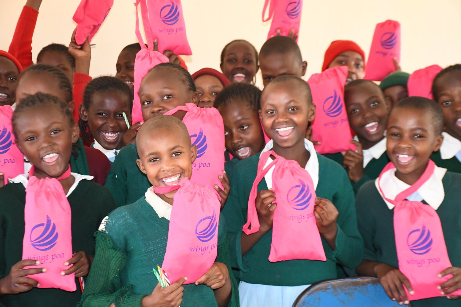 Children in green uniforms holding pink "Wings Poa" branded bags in an indoor setting.