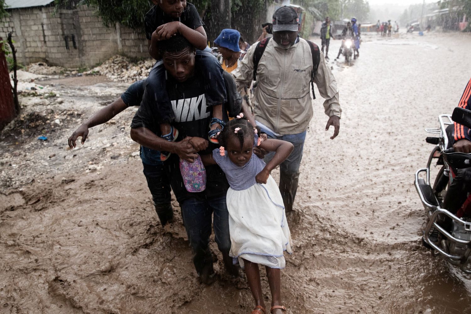 People navigate a muddy, rain-soaked street, with a man carrying a child on his shoulders and holding another child's hand.