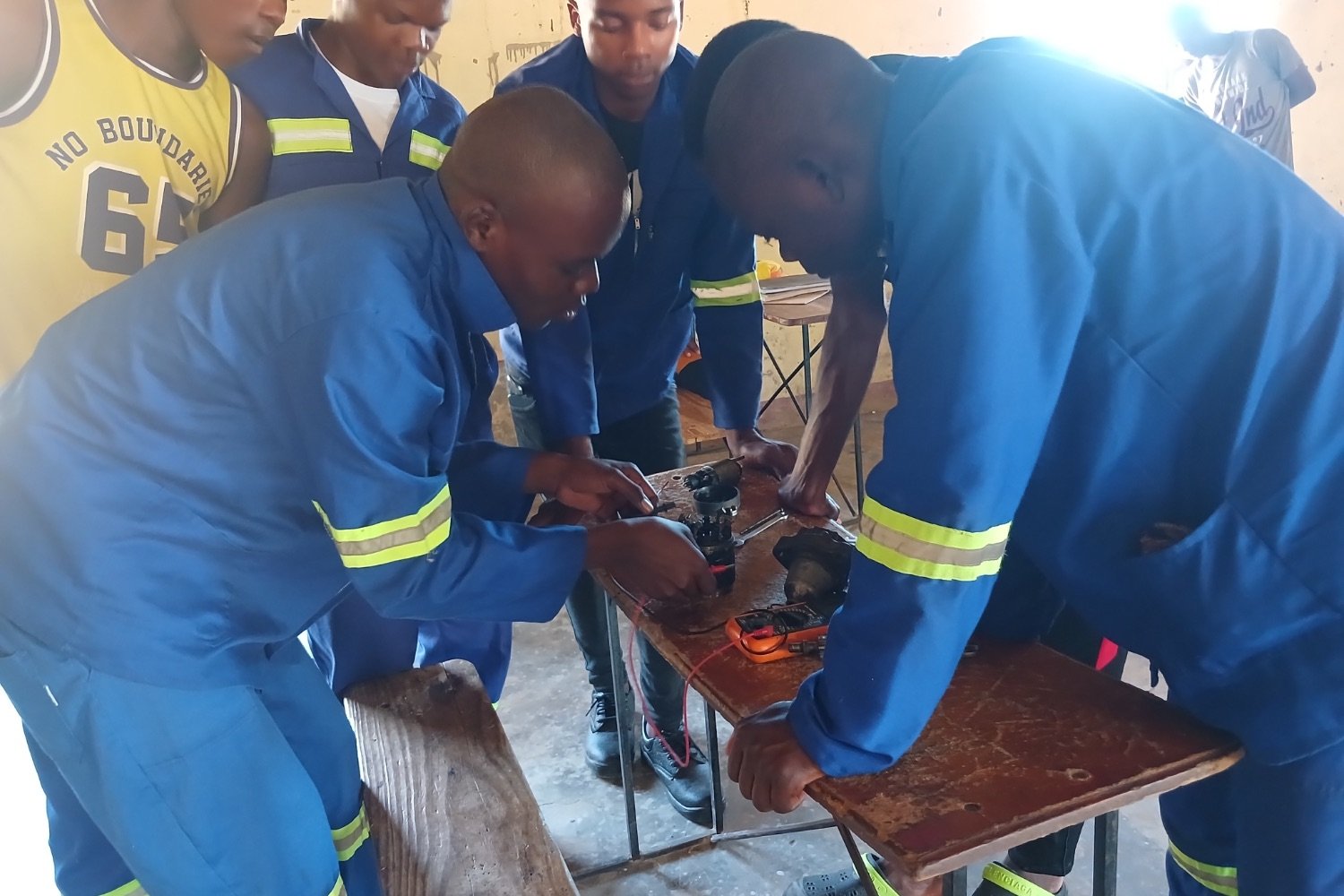 Students in blue reflective overalls working on an electrical wiring exercise on a worn wooden workbench in a vocational training classroom.