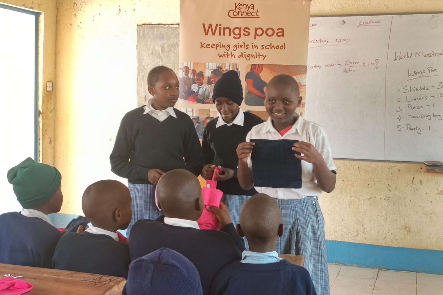 Girls in school uniforms presenting menstrual hygiene products in a Kenyan classroom with a Kenya Connect "Wings Poa" banner.
