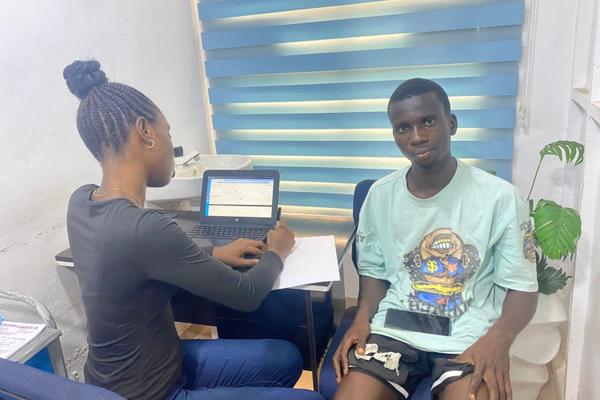 A young man in a mint green t-shirt sits opposite a woman using a laptop in a small clinic office in Lagos.