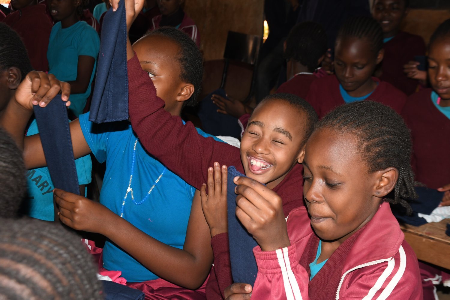 Children in maroon and blue uniforms holding dark blue fabric items inside a crowded classroom or hall.