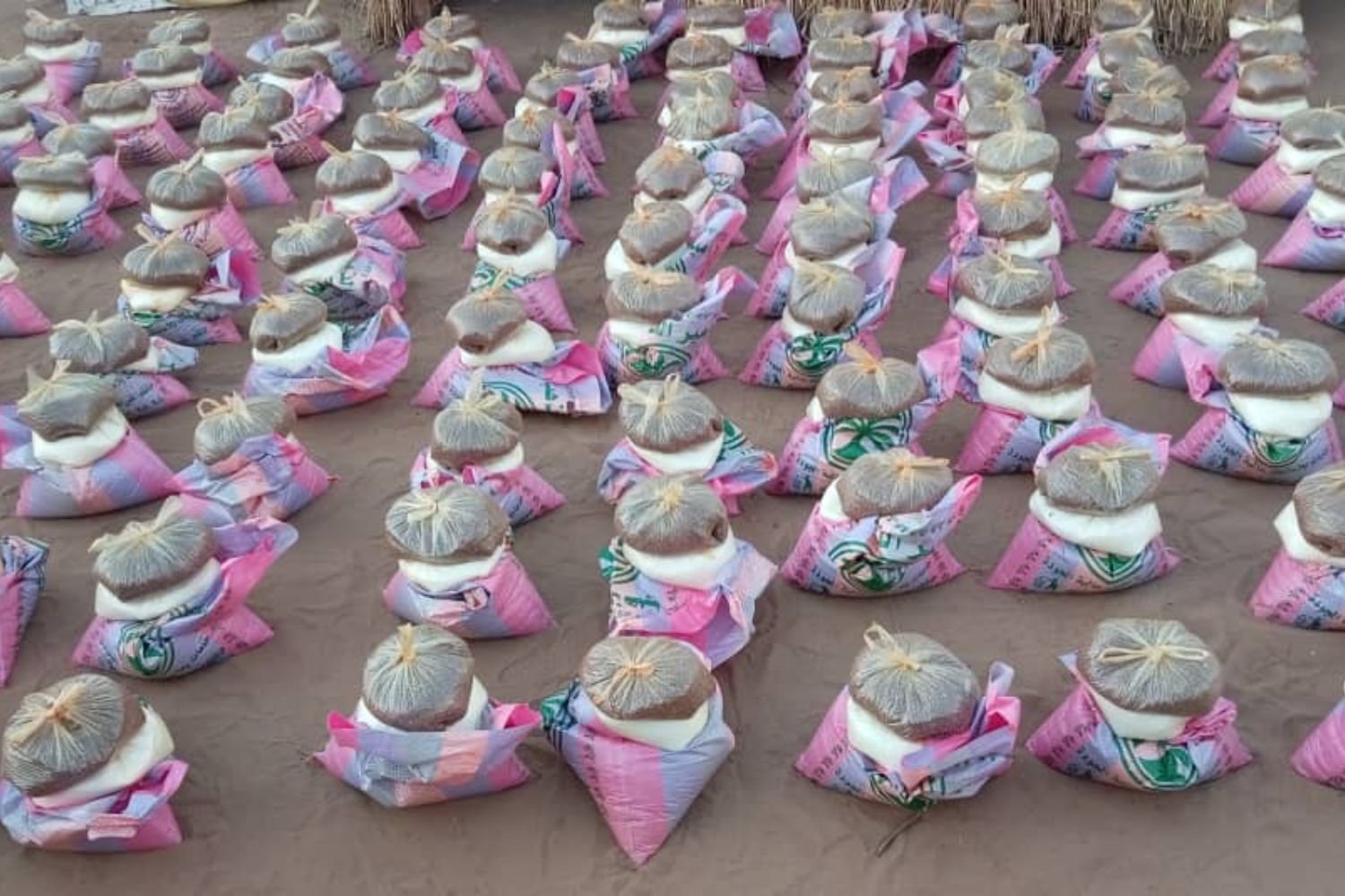Rows of pink fabric bags topped with brown bundled parcels arranged in a grid pattern on a sandy ground.