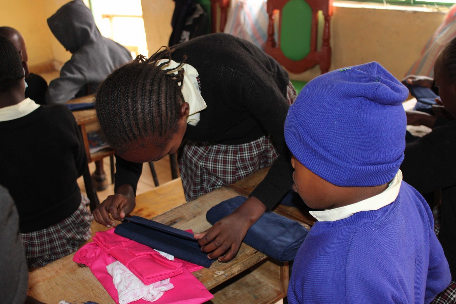 A girl in a black jumper and plaid skirt leans over a school desk examining folded clothing items beside a younger child in a blue hat.