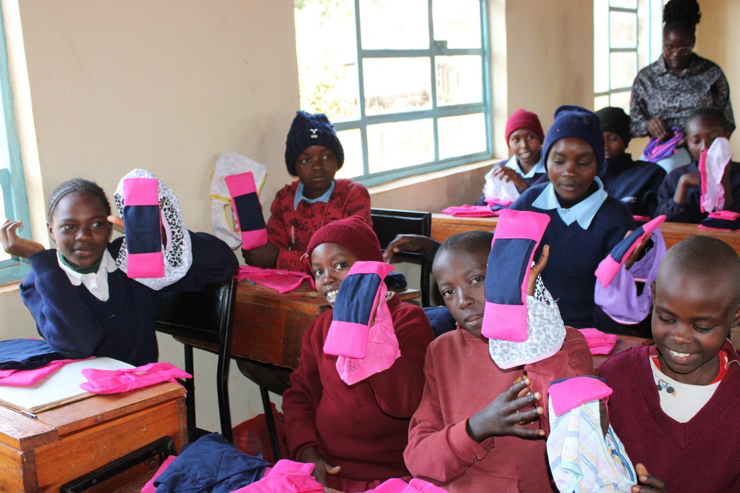Children in school uniforms holding pink and navy reusable sanitary pads in a classroom with wooden desks.