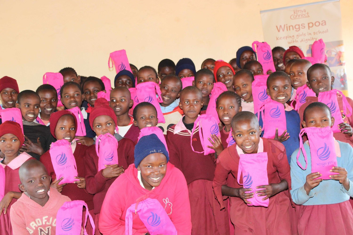 Girls holding pink "Wings Poa" drawstring bags gathered together indoors, with a Kenya Connect programme banner visible in the background.