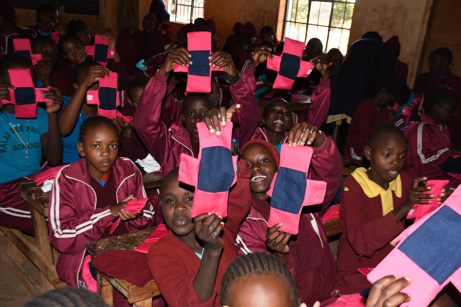 Children in maroon uniforms holding up pink and navy fabric items while seated at wooden desks in a classroom.
