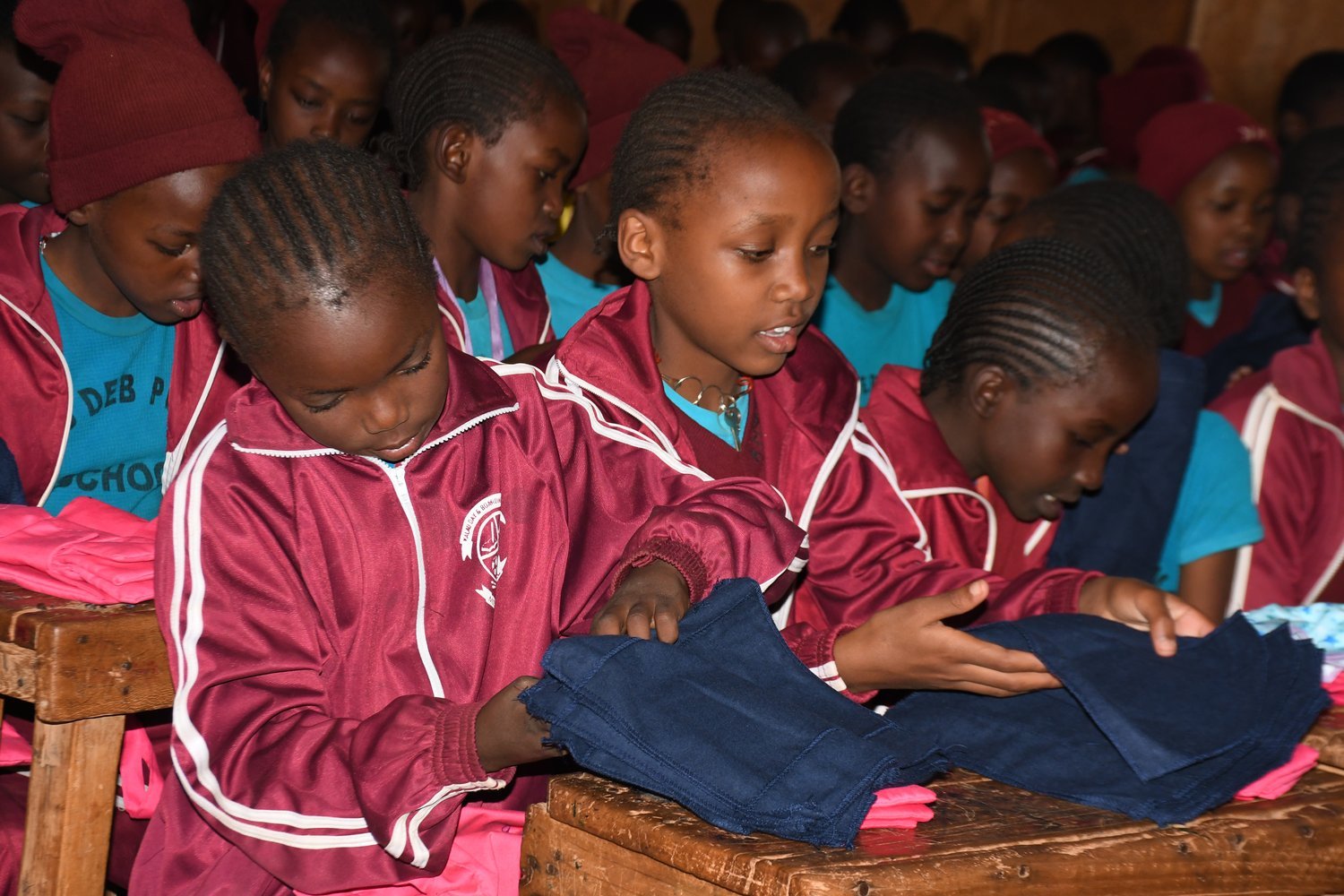 Girls in maroon school tracksuits examining navy blue clothing items at wooden desks in a crowded classroom.