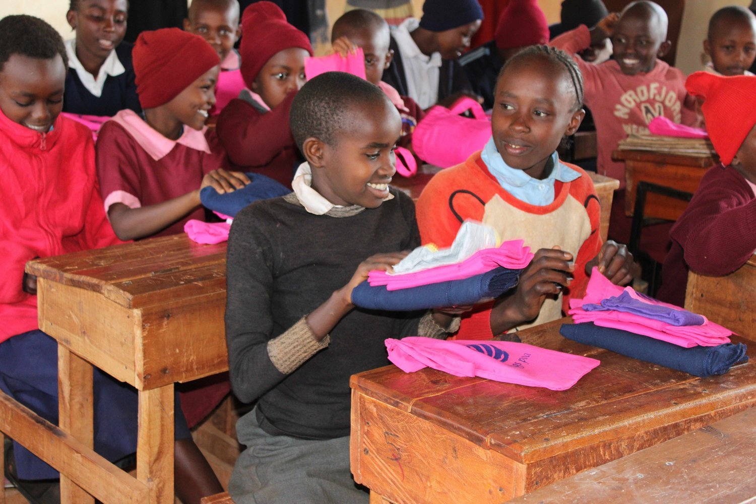 Children sitting at wooden desks holding pink and navy folded clothing items in a classroom.