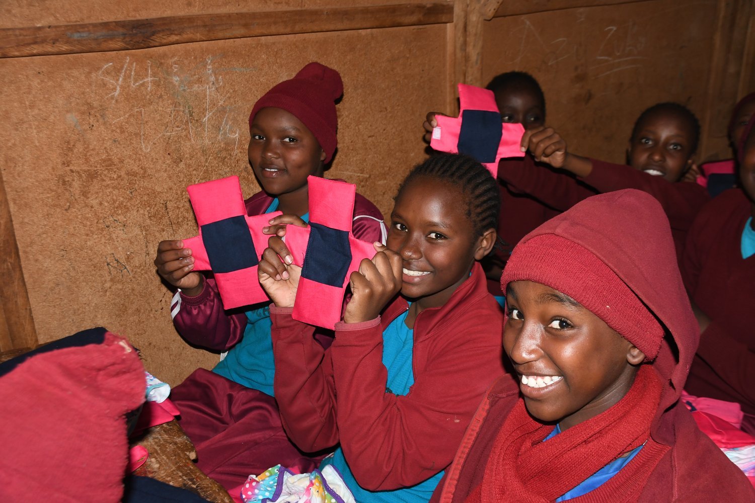 Girls in red uniforms holding pink and navy fabric items whilst seated inside a wooden classroom.
